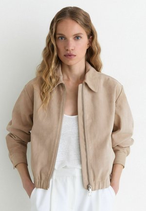 Junge Frau mit welligem blonden Haar trägt eine beige Reißverschlussjacke über einem weißen Top und steht vor einem schlichten weißen Hintergrund.