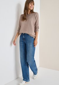 Beige gestreifter Pullover mit langen Ärmeln, kombiniert mit hellblauen weit geschnittenen Jeans und weißen Sneakers, vor einem neutralen Hintergrund stehend.