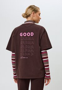Camiseta de algodón marrón con la frase "BUENA KARMA" impresa en texto rosa y negro en la parte posterior, con un corte relajado y mangas cortas.