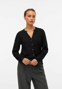 Chemise noire à manches longues, col rond, avec une petite poche poitrine ; fabriquée en tissu lisse avec un subtil éclat.