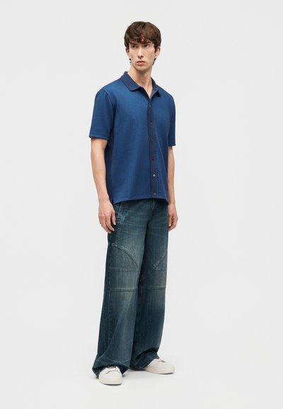 Jeune homme debout portant un polo bleu texturé, un jean en denim foncé à jambes larges avec des patchs, et des baskets blanches sur un fond uni.