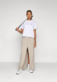 Bela kratka majica z logotipom, kombinirana z bež midi krilom s sprednjo zarezo. Model nosi bele superge in stoji pred nevtralnim ozadjem.