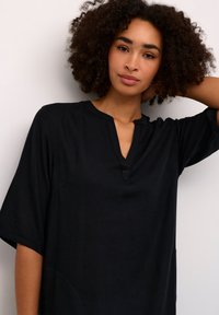 Black cotton top with a V-neck, short sleeves, and simple pocket detail. The fabric has a smooth, understated texture and classic design.