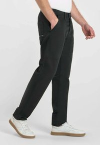 Man in zwarte rechte broek met zijrits en witte sneakers, die tegen een effen lichte achtergrond loopt.