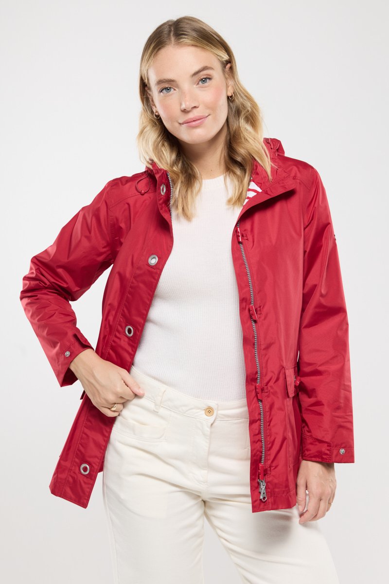Femme blonde portant un manteau de pluie rouge ouvert par-dessus une chemise blanche et un pantalon crème, se tenant avec une main dans sa poche.