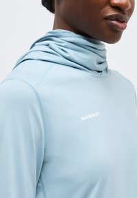 Približen posnetek osebe, ki nosi svetlo modro dolgotrajno majico Mammut z visokim ovratnikom, ki pokriva del brade in vratu.