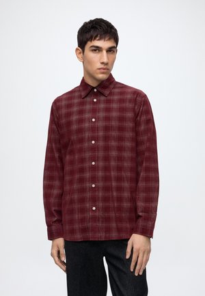 Homme portant une chemise à carreaux bordeaux à manches longues et un pantalon foncé, debout devant un fond clair uni.
