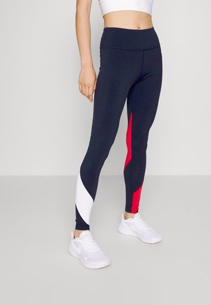 Leggings blu marina con accenti diagonali bianchi e rossi, caratterizzati da una vita alta e una texture liscia e elastica. Indossati con scarpe da ginnastica bianche.