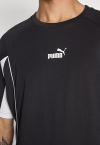 Černé sportovní tričko s bílými akcenty a logem Puma. Materiál má hladkou texturu a design s kulatým výstřihem, zdůrazňující sportovní styl.