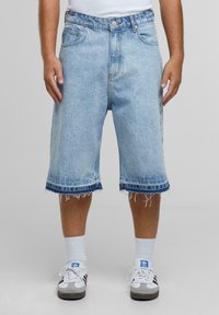 Pantalones cortos de mezclilla azul claro con un ajuste relajado, dobladillo deshilachado y costuras en azul oscuro contrastante. Combinados con calcetas blancas y zapatillas.