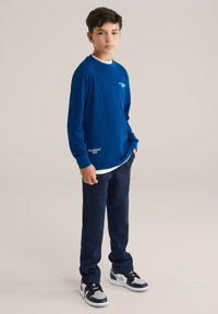 Modra srajca z dolgimi rokavi in teksturo, z belim logotipom, naložena čez belo srajco, v kombinaciji z mornarsko modrimi hlačami in črnimi supergami z sivo dodatki.