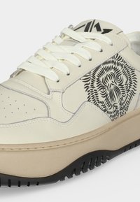 Sneaker color crema con lacci bianchi e suola nera, con un design dettagliato del volto di un leone nero sul pannello laterale.