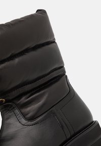 Botte noire en cuir montant à la cheville avec partie supérieure rembourrée et coutures visibles sur semelle lisse, vue de côté en gros plan.