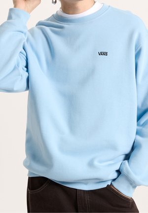 Personne portant un sweat-shirt Vans bleu clair par-dessus une chemise blanche, avec une main dans la poche d’un pantalon foncé et l’autre près du visage.