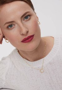 Złoty naszyjnik z okrągłym wisiorkiem, odsłonięty sweter w białym kolorze o fakturze dzianiny, kolczyki w kształcie kół, gładka skóra i odważny matowy kolor warg.