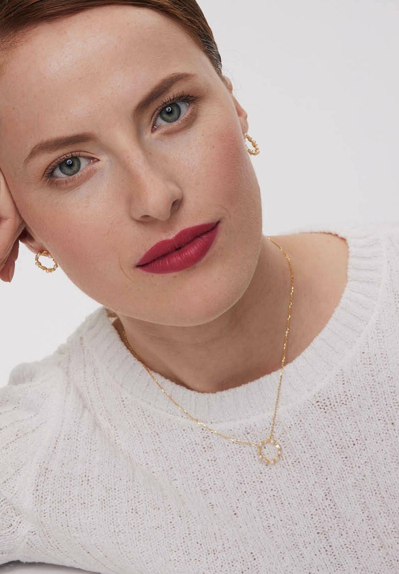 Złoty naszyjnik z okrągłym wisiorkiem, odsłonięty sweter w białym kolorze o fakturze dzianiny, kolczyki w kształcie kół, gładka skóra i odważny matowy kolor warg.