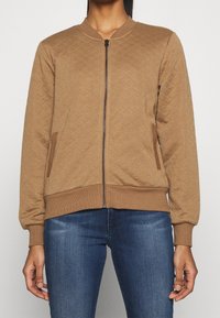 Veste bombardier matelassée marron avec fermeture éclair, poignets et ourlet côtelés, et poches latérales. Elle présente un tissu texturé avec un motif en losanges.