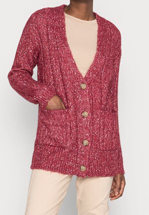 Persoon draagt een rood gebreid vest met knopen en voorzakken over een beige overhemd en broek, één hand rust in een zak.