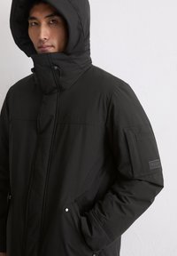 Schwarzer isolierter Mantel mit großer Kapuze, Reißverschluss, seitlichen Taschen und einem strukturierten Ärmelpatch. Glattes Material mit genähten Details.