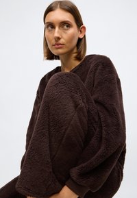 Brūns flīsa atpūtas apģērba komplekts ar teksturētu, ērti siltu apdari. Pārmērīgi liels tops ar kritiem pleciem un atbilstošas sašaurinātas bikses.