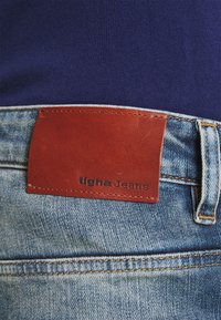 Dżinsy o jasnoniebieskim odcieniu, z brązową skórzaną łatą oznaczoną "tigha Jeans" w talii oraz widocznymi detalami przeszycia.