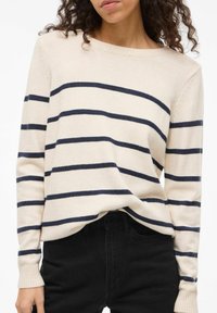 Sweter w kremowym kolorze z granatowymi poziomymi paskami, okrągłym dekoltem, ściągaczami na rękawach i luźnym krojem, zestawiony z czarnymi jeansami.