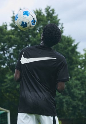 Młody mężczyzna kieruje piłką nożną głową na zewnątrz, ubrany w czarną koszulkę Nike i białe spodenki, z drzewami w tle.