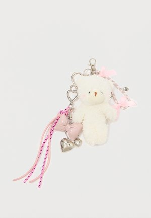 Avainkaulaketju, jossa on pehmeä valkoinen nalle, vaaleanpunainen solki, kaksi hopeista sydänkorua ja vaaleanpunaisia naruja mustilla yksityiskohdilla. Metallinen klipsi kiinnittämistä varten.