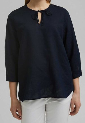 Frau trägt eine marineblaue Bluse mit langen Ärmeln, einer kleinen vorderen Schleife und weißen Hosen vor einfarbigem Hintergrund.