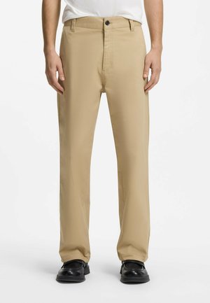 Mężczyzna w beżowych spodniach o prostych nogawkach z przednimi kieszeniami i czarnych butach, stojący na tle jednolitego białego tła.