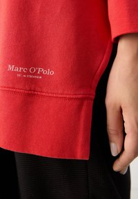 Rode sweatshirt met een gestructureerde afwerking, voorzien van een wit logo, naast een hand die rust tegen een zwart gestructureerd stof.