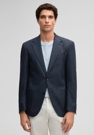 Homme portant un blazer bleu foncé sur une chemise bleu clair avec un pantalon beige, debout devant un fond gris uni.