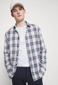 Camicia in flanella beige con motivo a quadri blu e grigio. Maniche lunghe, chiusura a bottoni, con tasca. Indossata sopra una maglia henley bianca.