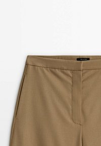 Pantalon beige taille haute avec fermeture éclair frontale cachée et poches latérales, présentant une texture de tissu lisse sur fond blanc.