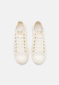 Białe trampki z płótna z złotymi oczkami, sznurowadłami i kolorowymi przeszyciami. Posiadają gumowe podeszwy i klasyczny design. Obecne logo Converse.