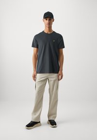 T-shirt à manches courtes gris foncé avec un petit logo jaune, associé à un pantalon cargo de couleur crème et des baskets noires, sur un fond uni.