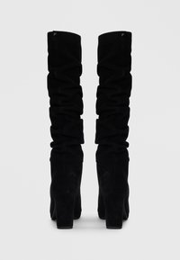 Bottes noires hautes en cuir suédé doux avec des détails froncés et des talons épais. Texture lisse avec une finition mate et un branding subtil.