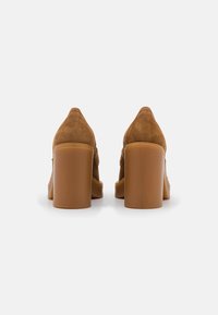 Brune ruskind højhælede sko med blokhæle og rund tå. Bagfra ses en glat tekstur og et minimalistisk design.