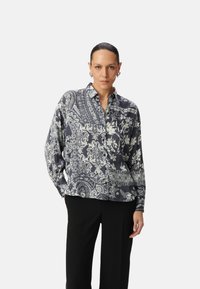Blusa a maniche lunghe di colore blu navy con intricati motivi floreali e paisley bianchi, apertura frontale con bottoni e vestibilità comoda. Indossata con pantaloni neri.