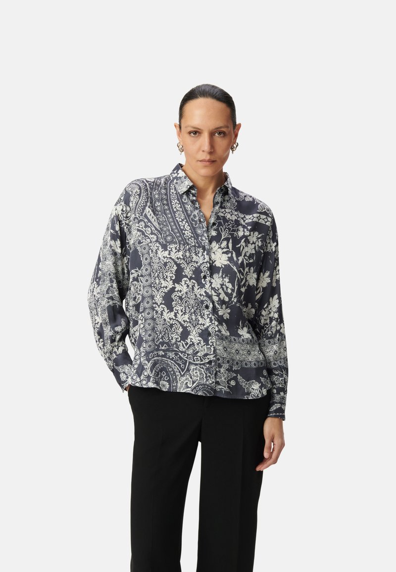 Blusa a maniche lunghe di colore blu navy con intricati motivi floreali e paisley bianchi, apertura frontale con bottoni e vestibilità comoda. Indossata con pantaloni neri.