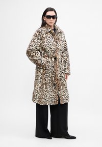 Kabát z imitácie kožuchu s leopardím vzorom a opaskom v páse, má špicatý golier, bočné vrecká a strednú dĺžku. Čierne nohavice a slnečné okuliare.