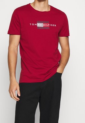 T-shirt en coton rouge avec un graphique rectangulaire affichant le texte "TOMMY HILFIGER" entouré de lignes horizontales blanches et grises. Manches courtes et col rond.