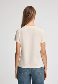 Ljusbeige t-shirt med kort ärm och rund halsringning, mjukt tyg, avslappnad passform och ren fåll; kombinerad med blå denimjeans.
