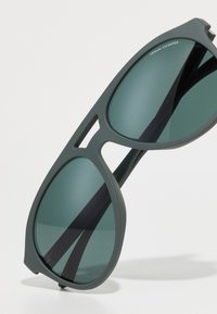 Ochelari de soare din plastic gri cu lentile mari, verzi, cu un design modern și accente decupate pe ramă. Logo-ul este vizibil pe lateral.