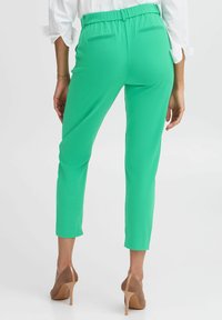 Pantalons verts, fuselés, avec une taille élastique et des poches arrière, associés à des chaussures à talons hauts nude. Texture de tissu lisse.