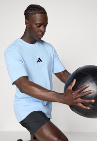 T-shirt azul de algodão com o logótipo preto da Adidas, combinado com calções pretos. O indivíduo segura uma bola medicinal preta com textura.