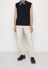 Suéter sin mangas de color azul marino, pantalones beiges, zapatos negros con suelas gruesas y una bufanda estampada alrededor del cuello. Diseño sencillo y ajustado.