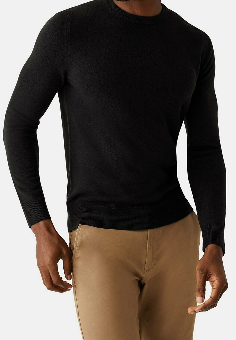 Pull en maille noire à manches longues, col rond et ourlet côtelé. Associé à un pantalon beige, mettant en valeur un tissu lisse et une coupe ajustée.