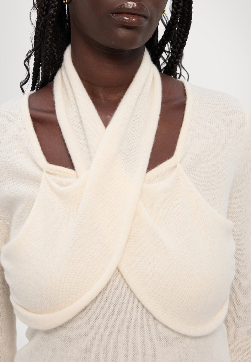 Pull en maille écru avec un design drapé unique, comportant des sections de tissu superposées et un col large et angulaire. La texture douce est visible.