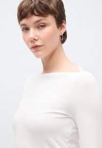 Fehér hosszú ujjú felső hajónyakú kivágással és puha textúrával, minimális varrással. A modell rövid hajú és egyszerű karika fülbevalót visel.